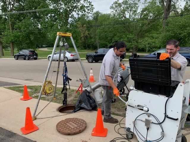 Two Trenchless Innovations technicians monitor equipment during a trenchless sewer line repair, working near an open manhole with safety cones and video inspection tools.