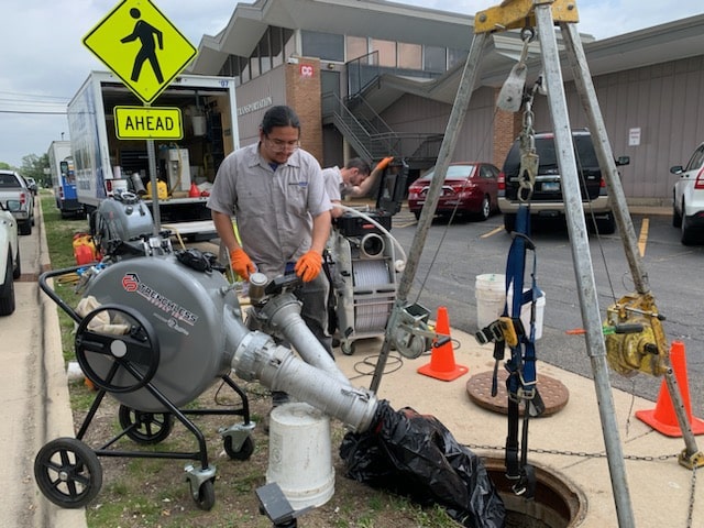 Trenchless Innovations technician operates sewer lining equipment outside a school facility near a manhole, using trenchless technology to repair underground pipes without excavation.